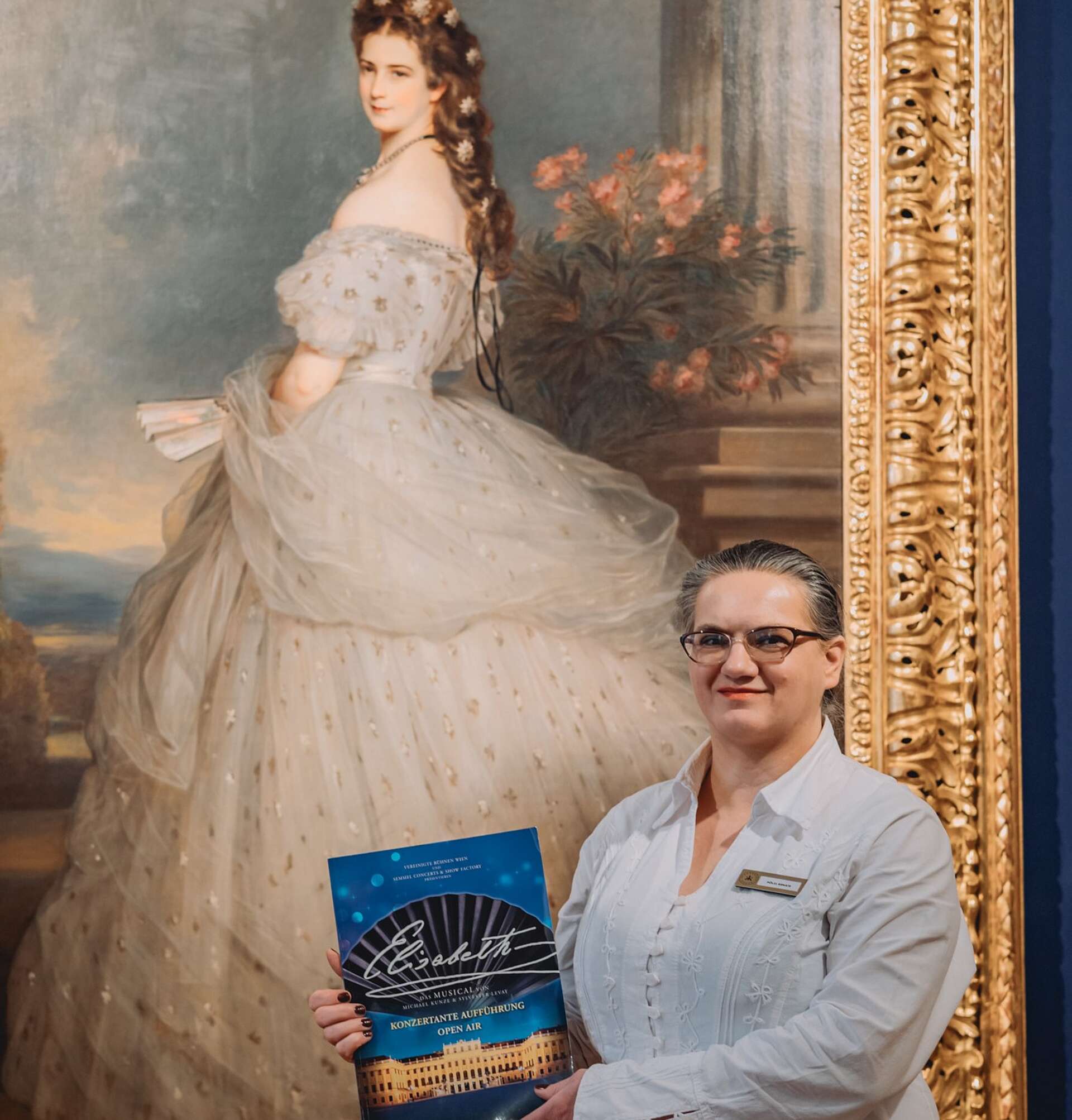 Mitarbeiterin hält das Elisabet Musical Programm hoch, während sie vor einem Portrait von Kaiserin Elisabeth steht